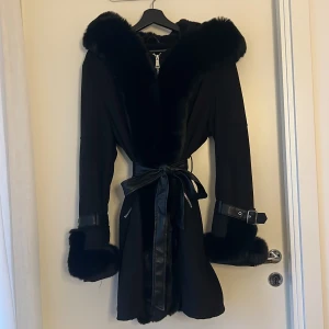 Svart pälsjacka från QED LDN med bälte - Snygg svart pälsjacka från QED LDN med stora fuskpälsdetaljer på krage och ärmslut. Jackan har bälte i midjan, dragkedja framtill och coola spännen vid ärmarna. Perfekt för dig som vill sticka ut med en lyxig vibe under kalla dagar.