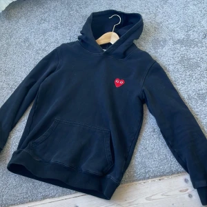Svart hoodie Comme des Garçons Play - Svart hoodie från Comme des Garçons Play med det ikoniska röda hjärtat med ögon broderat på bröstet. Tröjan har huva, känguruficka och långa ärmar. Mjuk bomullskvalitet som är skön att ha på sig. Perfekt för dig som gillar streetwear och vill ha något stilrent med en twist.