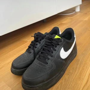 Nike Air Force 1 svart/vit sneakers - Svarta Nike Air Force 1 sneakers med vit swoosh och detaljer i neongult på plösen och hälen. Klassisk låg modell med svart snörning och perforerad tå. Ovandel i skinn med platt sula och rund tå. Perfekta för dig som gillar streetstyle.