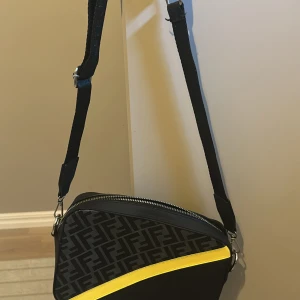 Fendi axelväska med FF-mönster och gul detalj - Säljer en svart axelväska från Fendi med klassiskt FF-mönster på framsidan och en snygg gul diagonal detalj. Väskan har justerbar axelrem, dragkedja upptill och kombinerar skinn och textil för en modern vibe.