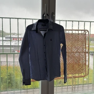 Blå prickig skjorta Polo Ralph Lauren - Snygg blå skjorta från Polo Ralph Lauren med små vita prickar och klassisk krage. Långärmad modell med svarta knappar och diskret logga på bröstet. Perfekt för dig som gillar stilrena detaljer och tidlös design.