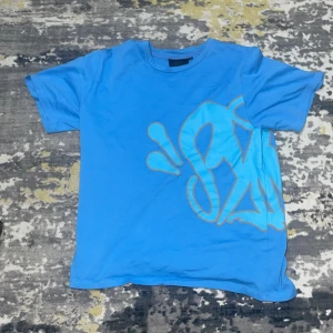 Blå t-shirt med graffiti från OX Fuckeries - Blå t-shirt från OX Fuckeries i storlek S med stort graffiti-inspirerat tryck i ljusblått och grått över både fram- och baksida. Skön passform och klassisk rund hals. Perfekt för dig som gillar streetwear och unika prints.