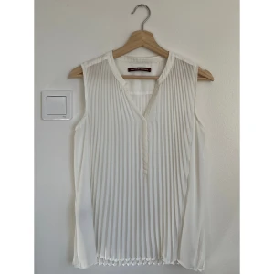 Vit plisserad blus Comptoir des Cotonniers - En snygg vit blus från Comptoir des Cotonniers. Blusen är ärmlös med v-ringning och har plisserat tyg framtill och baktill. Tunt, lätt material som ger ett luftigt intryck. Perfekt för dig som gillar stilrena och eleganta plagg.