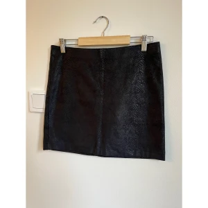 Svart kortkjol i mockaimitation från Promod - Snygg svart kortkjol från Promod med diskret ormskinnsmönster. Kjolen har elastisk midja. Perfekt för dig som gillar stilrena detaljer med lite edge.