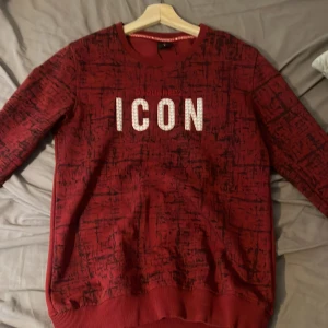 Röd Dsquared2 ICON sweatshirt S - Röd sweatshirt från Dsquared2 med svart abstrakt mönster på framsidan och ärmarna. Stor vit 'ICON'-text med små röda detaljer på bröstet. Rund halsringning och ribbade muddar. Perfekt för dig som vill sticka ut med en streetstyle-look.