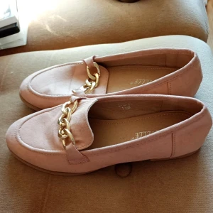 Rosa loafers med guldkedja - Snygga rosa loafers i mocka-look med en chunky guldfärgad kedjedetalj framtill. Skorna har platt sula och rund tå, perfekta för en trendig och avslappnad stil. Passar till både jeans och kjol för en cool vardagslook.