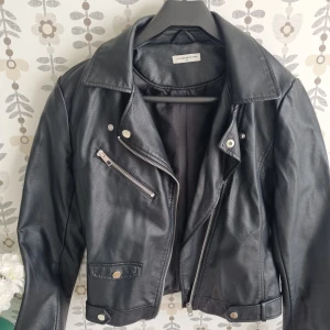 Svart skinnjacka från Jacqueline de Yong - Snygg svart skinnjacka med klassisk bikerstil från Jacqueline de Yong. Jackan har silvriga dragkedjor, knappar och detaljer, samt en croppad passform och bred krage. Perfekt för dig som vill ha en edgy look med attityd.