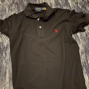 Svart Polo Ralph Lauren t-shirt - Svart t-shirt från Polo Ralph Lauren med klassisk krage och två knappar framtill. Custom slim fit-modell med rött broderat Polo-logo på bröstet. Tillverkad i mjuk bomull som känns skön mot huden. Perfekt för dig som gillar stilren och enkel design. PRIS KAN DISKUTERAS VID SNABB KÖP!!!
