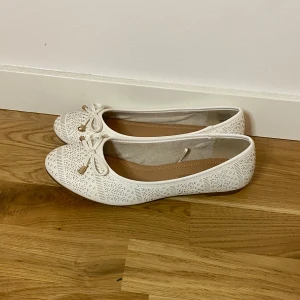 Vita ballerinaskor med rosett - Snygga vita ballerinaskor med rund tå och dekorativ rosett framtill. Skorna har ett coolt geometriskt mönster och små metalldetaljer på rosetten. Perfekt för dig som gillar stilrena och trendiga skor med lite extra detaljer.