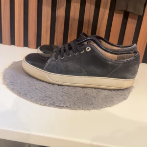 Svarta sneakers med mockadetaljer - Säljer ett par svarta Lanvins med snygga mockadetaljer och blank tå i syntet. Skorna har snörning med metallöljetter och en vit platt sula som ger en klassisk look. Perfekta för dig som gillar stilrena och enkla sneakers med en twist. Tvättar dem innan jag skickar 