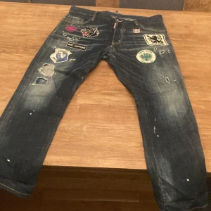 DSQUARED2 jeans med patches, strl 50 - Unika blå jeans från DSQUARED2 med coola patches och slitningar framtill och baktill. Jeansen har raka ben, broderade detaljer och klassisk femficksmodell. Snygg tvätt och trendiga slitningar ger en edgy vibe. Perfekta för dig som vill sticka ut.