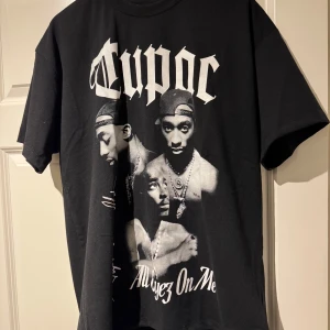 Svart Tupac t-shirt Pull&Bear L - Svart t-shirt från Pull&Bear med stort Tupac-tryck och texten 'All Eyez On Me' på framsidan. Klassisk rund hals och korta ärmar. Materialet är mjuk bomull och passformen är normal. Perfekt för dig som gillar streetwear och ikoniska musikmotiv.