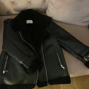 Svart teddyjacka från Stradivarius - Snygg svart teddyjacka från Stradivarius i storlek L. Jackan har mjukt foder, bred krage, flera silvriga dragkedjor och coola spänndetaljer vid ärmsluten. Perfekt för dig som vill ha en stilren och trendig look under kyliga dagar.