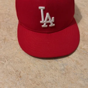 Röd LA 59FIFTY keps från New Era - Snygg röd 59FIFTY keps från New Era med LA-logga broderad i vitt framtill. Klassisk flat brim och MLB-märke baktill. Kepsen är i storlek 7 3/8 och har en stilren look som passar perfekt till streetwear. Materialet är bomull och kepsen har en strukturerad form.