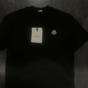 Svart Moncler t-shirt med logga - Svart t-shirt från Moncler med klassisk rund hals och korta ärmar. På bröstet sitter Monclers ikoniska logopatch i vitt och rött. T-shirten är gjord i mjuk bomull och har en clean, stilren look som funkar till allt. Perfekt för dig som gillar exklusiva märken.