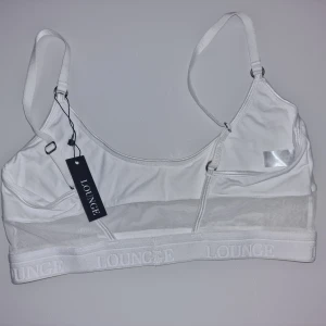Vit mesh bh från Lounge - Vit bh från Lounge med justerbara axelband och bred resår med logga. Toppen har en snygg meshpanel under bysten som ger en sportig vibe. För dig som gillar stilrena och enkla plagg.