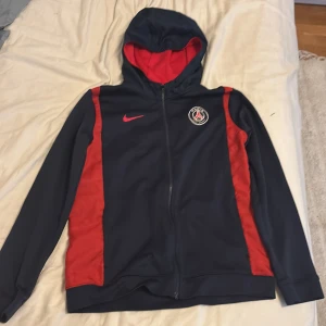 PSG hoodie från Nike 158-170  - Mörkblå hoodie från Nike i storlek XL med Paris Saint-Germain-logga på bröstet och röda detaljer längs sidorna. Tröjan har hel dragkedja, huva med röd insida och långa ärmar. Perfekt för dig som gillar fotboll och vill ha en sportig look.