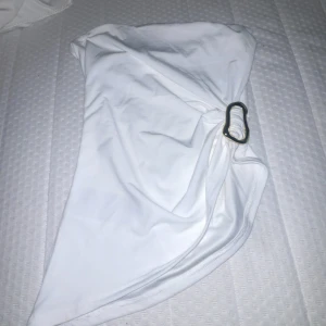 Vit topp med en guldiga metall grej  - Snygg vit topp med en cool detalj i form av en metallring på sidan. Toppen har en enkel och stilren design med mjukt syntetmaterial som känns skönt mot huden.