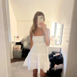 Vit kort sommarklänning  - Supersöt vit klänning från Neworker med smock upptill och tunna axelband. Kjolen är volangig och har diskreta ränder i tyget. Perfekt för varma dagar och har en lätt, luftig känsla. Helt oanvänd med prislapp kvar.