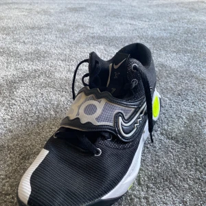 Svarta Nike sneakers med neon och vitt - Snygga svarta Nike sneakers med vita och neongula detaljer. Skorna har en cool design med snörning, bred kardborre över foten och tydlig Nike-logga på sidan. Ovandelen är i syntetmaterial och mesh för bra andningsförmåga. Perfekta för dig som gillar sportig stil.