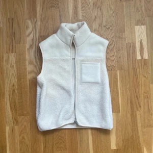 Vit Teddy väst - Mysig vit fleeceväst med hög krage och dragkedja framtill. Västen har en enkel design med en liten bröstficka på vänster sida och är tillverkad i mjukt, fluffigt fleecematerial. Perfekt att slänga på sig över en hoodie eller långärmad tröja.