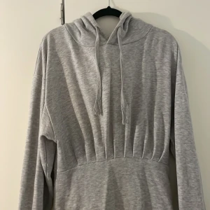 Grå lång hoodieklänning med slits - Mysig grå långklänning i sweatshirtmaterial med huva och dragsko. Klänningen har långa ärmar med muddar, markerad midja och en slits bak för extra rörelsefrihet. Perfekt för en chill och avslappnad stil.