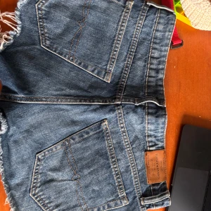 Jeansshorts från Lucky Brand - Snygga blå jeansshorts från Lucky Brand med fransig kant och klassiska fickor både fram och bak. Shortsen har normal passform och är perfekta för sommaren. De är tillverkade i slitstarkt denim och har en cool, avslappnad vibe.