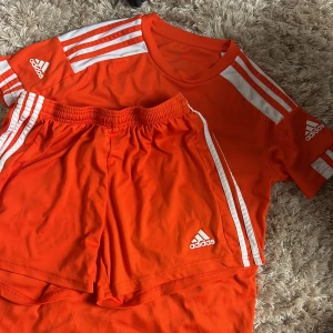 Orangea Adidas shorts och T-shirt med vita ränder - Snygga orangea träningsshorts och T-shirt från Adidas med klassiska vita ränder längs sidorna och Adidas-logga framtill. Shortsen har elastisk midja och är gjorda i ett lätt och svalt polyestermaterial, perfekta för sport eller träning.