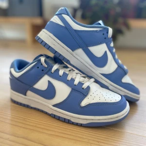 Nike Dunk Low blå och vita sneakers - Nike Dunk Low sneakers i fräscha blå och vita färger. Skorna har klassisk snörning, perforerad tå och ikonisk Swoosh på sidorna. Ovandel i skinn med vit bas och blå overlays, samt matchande blå yttersula. Perfekta för dig som gillar streetwear och vill sticka ut.