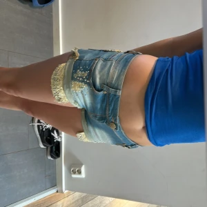 Blå jeansshorts med pärlor och spets - Snygga blå jeansshorts från Monday Denim med råa kanter och slitningar. Dekorerade med gul spets och glittrande pärlor på fickor och framsida. Shortsen har klassisk femficksdesign, knappstängning och coola detaljer som gör dem unika.