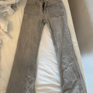 Grå bootcut jeans med fickdetaljer - Snygga grå jeans med bootcut passform och coola fickor bak med lock och metallknappar. Jeansen har klassisk femficksdesign och är tillverkade i mjukt denimtyg. Perfekta för dig som gillar en avslappnad men trendig stil.
