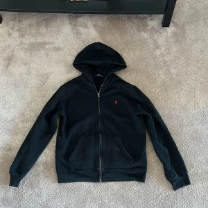 Svart hoodie från Ralph Lauren - Svart hoodie från Ralph Lauren med dragkedja och klassisk huva. Tröjan har en röd broderad logga på bröstet och två stora fickor framtill. 