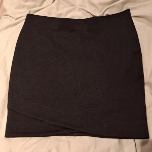 Svart omlottkjol från H&M - Snygg svart kortkjol från H&M i storlek 38. Kjolen har en omlott-detalj framtill. Materialet är mjukt och stretchigt.. Passformen är normal och kjolen är enkel att matcha med olika toppar.