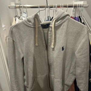 Grå hoodie från Polo Ralph Lauren - Säljer en grå hoodie från Polo Ralph Lauren med dragkedja och huva med snörning. Klassisk blå logga framtill. Perfekt för chill dagar. Fråga gärna om fler bilder och förslå gärna ditt pris!