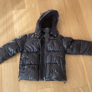 Svart pufferjacka från Nelly, M - Snygg svart pufferjacka från Nelly med glansig finish och huva. Jackan har dragkedja framtill, två fickor och quiltad design som ger en cool vibe. Perfekt för kalla dagar när du vill hålla stilen och värmen.