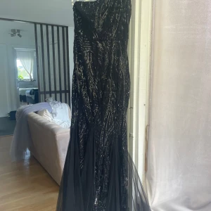 Svart glittrig långklänning med paljetter - Säljer en svart långklänning med glittriga paljetter och mönstrad yta. Klänningen har en elegant, figurnära siluett, stretchig och bekväm tyg och har en axelband med ett svepande tyllparti nedtill. Perfekt för bal eller fest när du vill sticka ut med en glam look. Använd endast en gång. Mått: Storlek M men kan passa som L, Längd 154cm från axelband till nedersta. Tyg: 95% Polyester