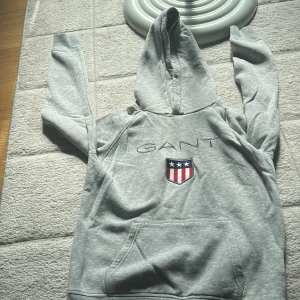 Grå hoodie från GANT med brodyr - Snygg  grå hoodie från GANT med broderad logga och sköld på bröstet. Tröjan har huva, magficka och klassiska ränder i rött, vitt och blått på insidan av kragen. Perfekt för en chill och sportig look. Använd endast en gång fick den som present och är för storlek 152-158 och nypris 799