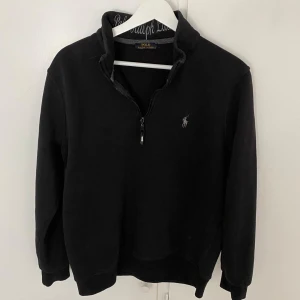 Svart half zip tröja från Polo Ralph Lauren - Svart långärmad tröja från Polo Ralph Lauren med half zip och broderad logga på bröstet. Klassisk passform med hög krage och ribbade muddar. Perfekt för lager-på-lager och chill stil. Materialet är mjuk bomullsmix.