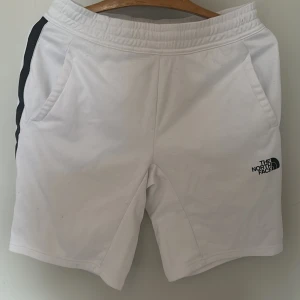 Vita shorts från The North Face - Snygga vita shorts från The North Face med svart logga på benet och svarta detaljer längs sidorna. Shortsen har elastisk midja och sidofickor, samt en cool rand med The North Face-tryck längs ena sidan. Perfekta för sommaren och träning.
