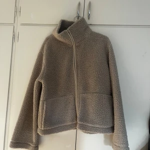 Beige teddyjacka från 157, stl M - Supermjuk och mysig beige teddyjacka från 157 med hög krage och dragkedja framtill. Jackan har två stora fickor och är perfekt för kyliga dagar. Oversized fit för en chill vibe.
