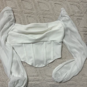 Vit korsettopp med puffärmar - Supersnygg vit korsettopp med puffiga långärmar i transparent tyg. Toppen har markerade sömmar framtill och en snygg draperad halsringning. Perfekt för dig som vill sticka ut med en trendig och feminin look.