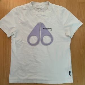 Vit Moose Knuckles t-shirt med  print - Snygg vit t-shirt från Moose Knuckles med ett stort  logoprint på bröstet och texten 'Moose Knuckles Engineered in Canada'. På ryggen finns ett  'Moose'-tryck. Klassisk passform och rund hals. Tillverkad i mjuk 100% bomull