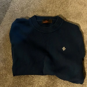 Mörkblå stickad tröja från Morris - Snygg mörkblå stickad tröja från Morris med rund halsringning och diskret logga broderad på bröstet. Tröjan har ribbade muddar vid ärmslut och nederkant, perfekt för dig som gillar klassisk och stilren look.