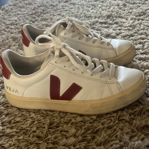 Vita Veja sneakers med röda detaljer - Säljer ett par klassiska Veja sneakers i vitt skinn med röda detaljer och logga på sidan och hälen. Modellen har rund tå, platt sula och snörning framtill. Perfekt för dig som gillar clean stil med en touch av färg. Storlek 38.