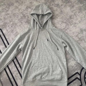 Grå hoodie från Polo Ralph Lauren - Snygg grå hoodie från Polo Ralph Lauren med klassisk broderad logga på bröstet. Tröjan har huva med snörning, känguruficka och långa ärmar. Perfekt för en chill och stilren look.