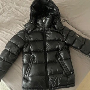 Svart Moncler pufferjacka med huva - Säljer en svart pufferjacka från Moncler med glansig finish, huva och dragkedja framtill. Jackan har två snedställda fickor med dragkedja och en ärmficka med Moncler-logga. Perfekt för kalla dagar och riktigt snygg. Storlek 00. Har inga kvitton eller något liknande som visar att den är äkta. Vet att jag var med när min vän köpte jackan från butiken. Sedan köpte jag den från henne men då hade hon inte kvar något kvitto. Pris kan diskuteras vid snabbt köp!!!
