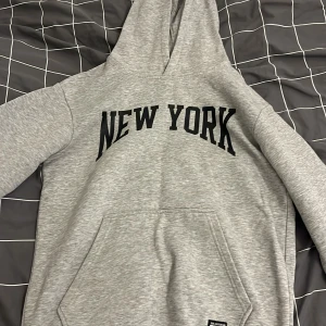 Grå hoodie med New York-tryck - Snygg grå hoodie med svart 'NEW YORK'-tryck över bröstet. Klassisk känguruficka framtill och stor huva. Perfekt för chill dagar eller när du vill ha en enkel streetstyle-look. Mjuk och skön bomullsblandning, långärmad och normal passform.