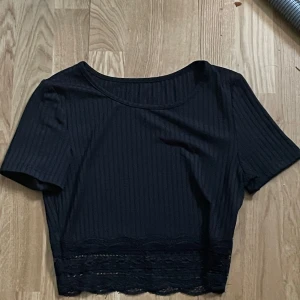 Svart croppad ribbad topp med spets - Svart croppad topp med ribbad struktur och korta ärmar. Toppen har en rund halsringning och en snygg spetskant längst ner som ger en extra detalj. Perfekt för att lyfta vilken outfit som helst.