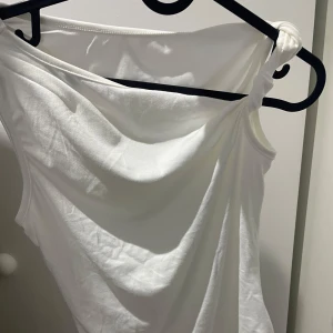 Vit oneshoulder topp i bomull - Säljer en vit oneshoulder topp med snygg drapering och knutdetalj på axeln. Toppen är ärmlös och har en clean, stilren look som passar perfekt till jeans eller kjol. Gjord i mjuk bomull för en skön känsla hela dagen. Från shein därav priset.