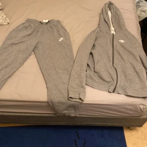 Grå Nike dress tröja och byxor  - Grå tröja från Nike med luva och dragkedja framtill. Klassisk sportig look med vit Nike-logga på bröstet. Tröjan är i mjukt material och har fickor på sidorna. Perfekt för en chill och avslappnad stil. Tröjan och byxorna kostar 200 tillsammans! Storlek är från 158 cm till 170cm